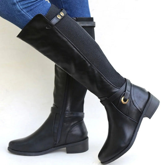 Black knee-high riding boots with wide ribbed stretch side panels, ankle straps with metal hardware, rounded square toes, and low block heels, shown worn with jeans.