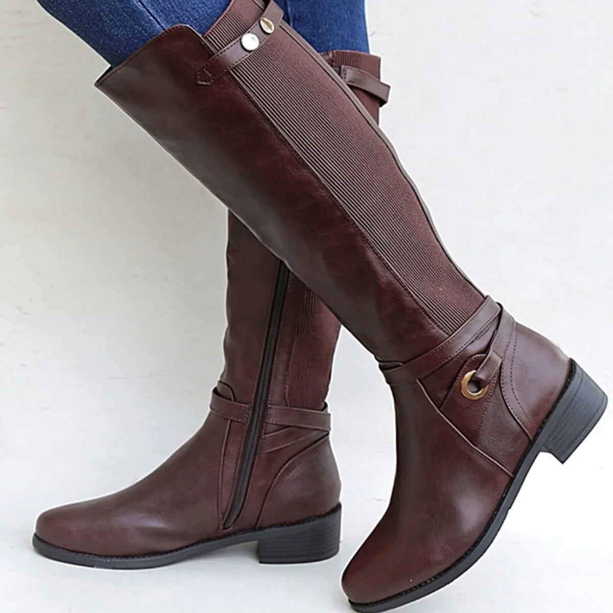 Brown knee-high riding boots featuring contrasting ribbed stretch shaft panels, decorative ankle straps, metal accents, rounded square toes, and low block heels.