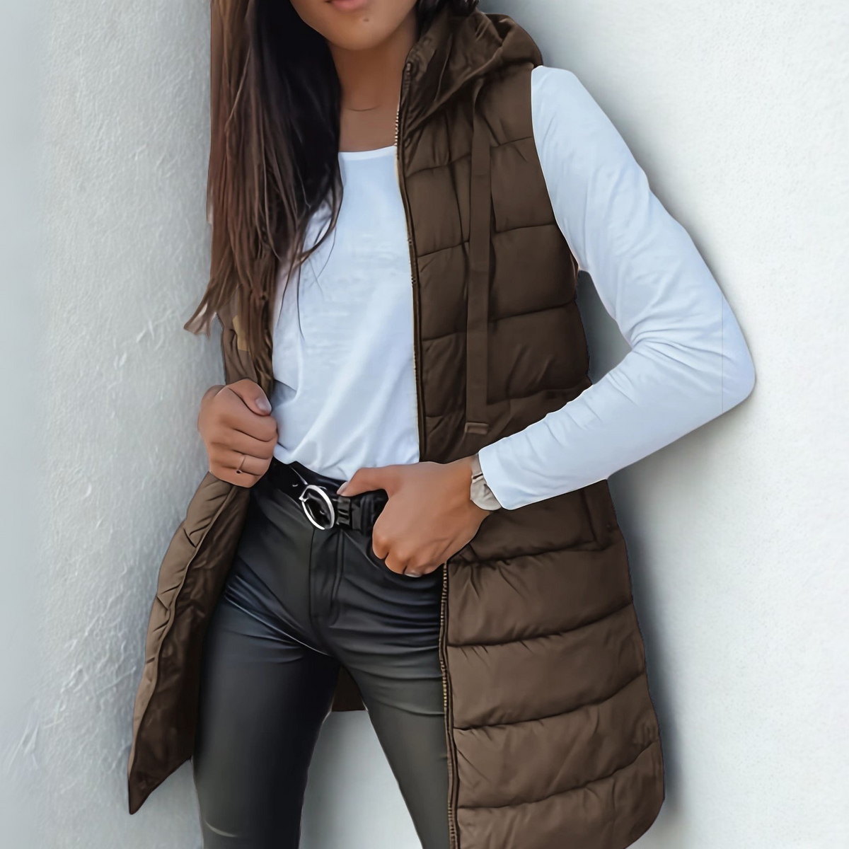 Dark brown long puffer vest styled over a white shirt, with its attached hood, open front zipper, and horizontal quilted sections visible.