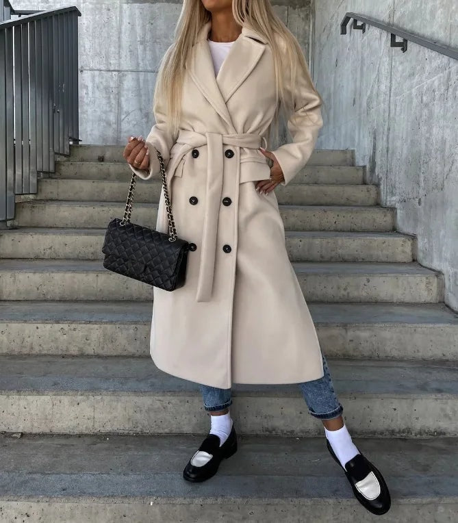 Light beige longline coat with notched lapels, double rows of buttons, and front flap pockets, styled with rolled jeans.