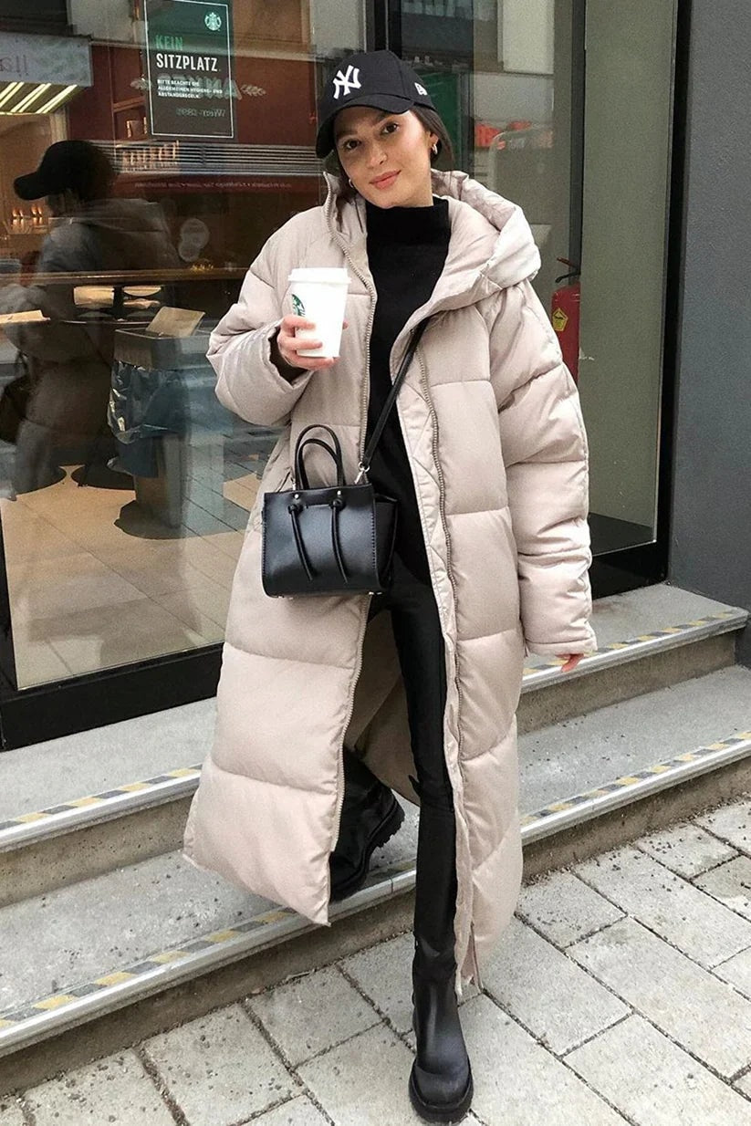 Light-colored longline hooded puffer coat worn open over black layers with a crossbody handbag and boots, showing its extended length and relaxed fit.