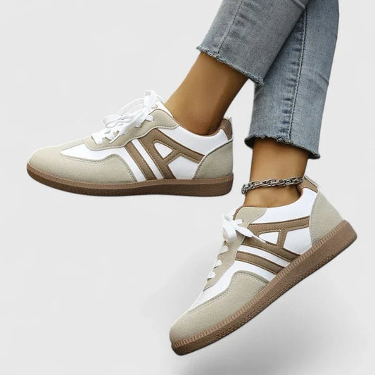 Low-top lace-up sneakers shown in a side view, featuring light neutral panels, white accents, layered contrast side detailing, and brown textured outsoles.