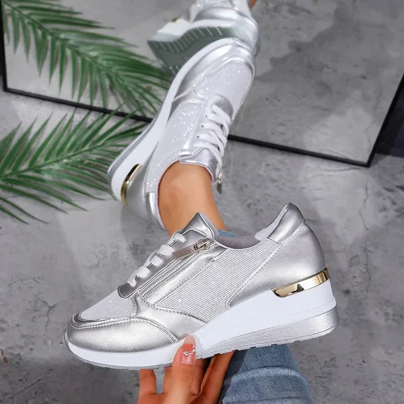 Silver metallic sneaker held in profile, displaying the low-top shape, side zip, platform sole, and gold-toned heel detail.