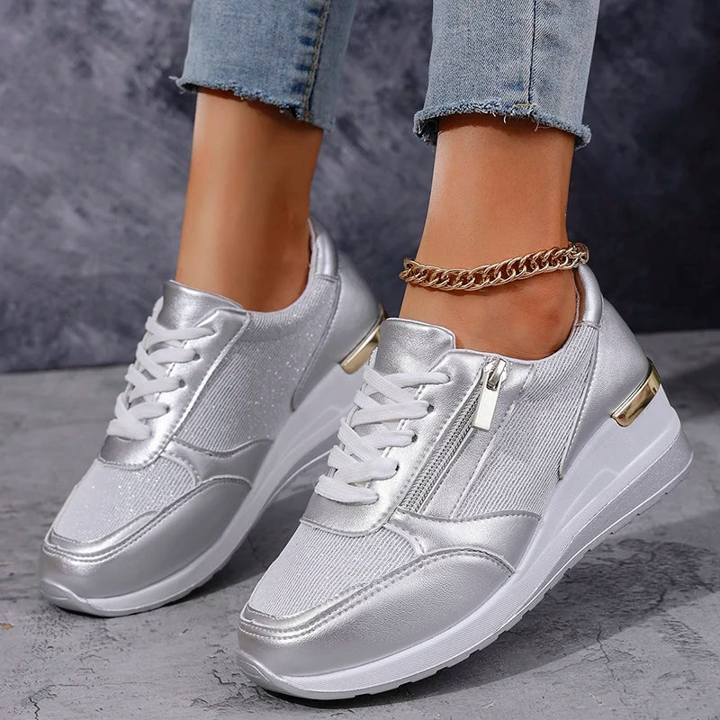 Pair of silver metallic platform sneakers angled toward the viewer, with white laces, visible side zips, and gold-toned heel accents.