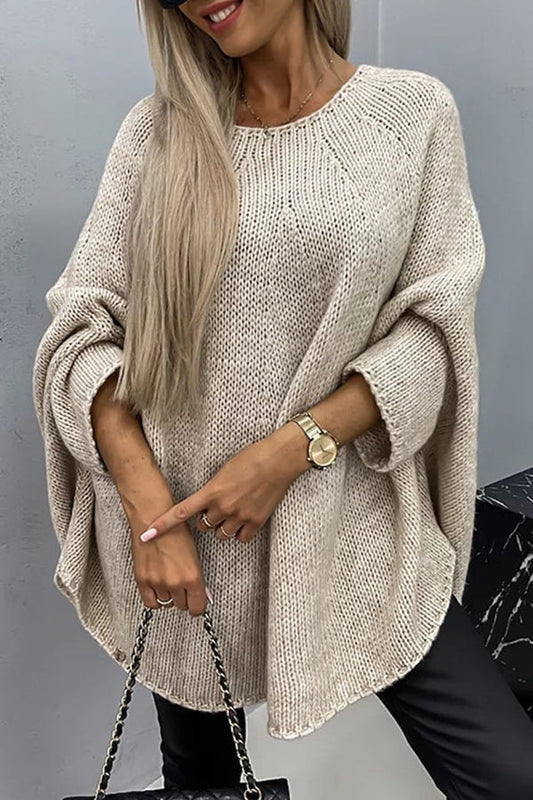 Beige oversized knit poncho sweater with a crew neck, wide sleeves, open knit texture, and curved tunic-length hem styled with black trousers.