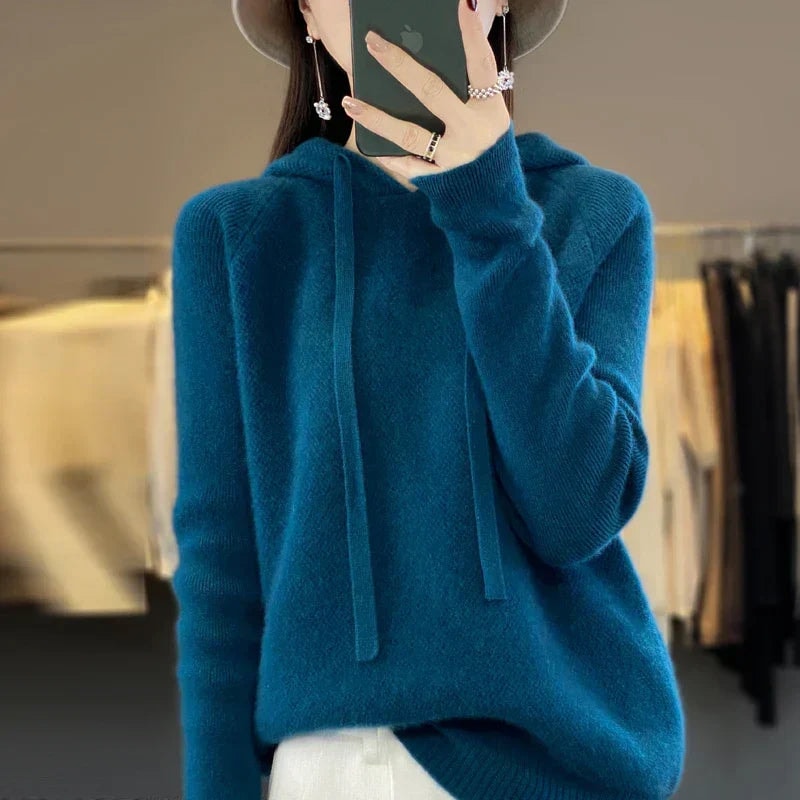 Deep teal oversized knit pullover hoodie with a hood and long drawstrings, styled over light trousers.