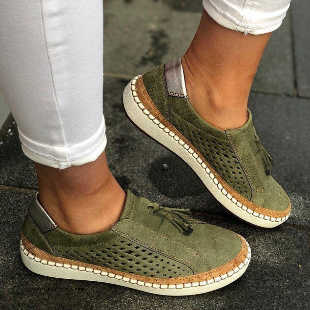 Olive green perforated slip-on sneakers with decorative front tassels and contrasting stitched soles, shown worn with white cropped jeans.