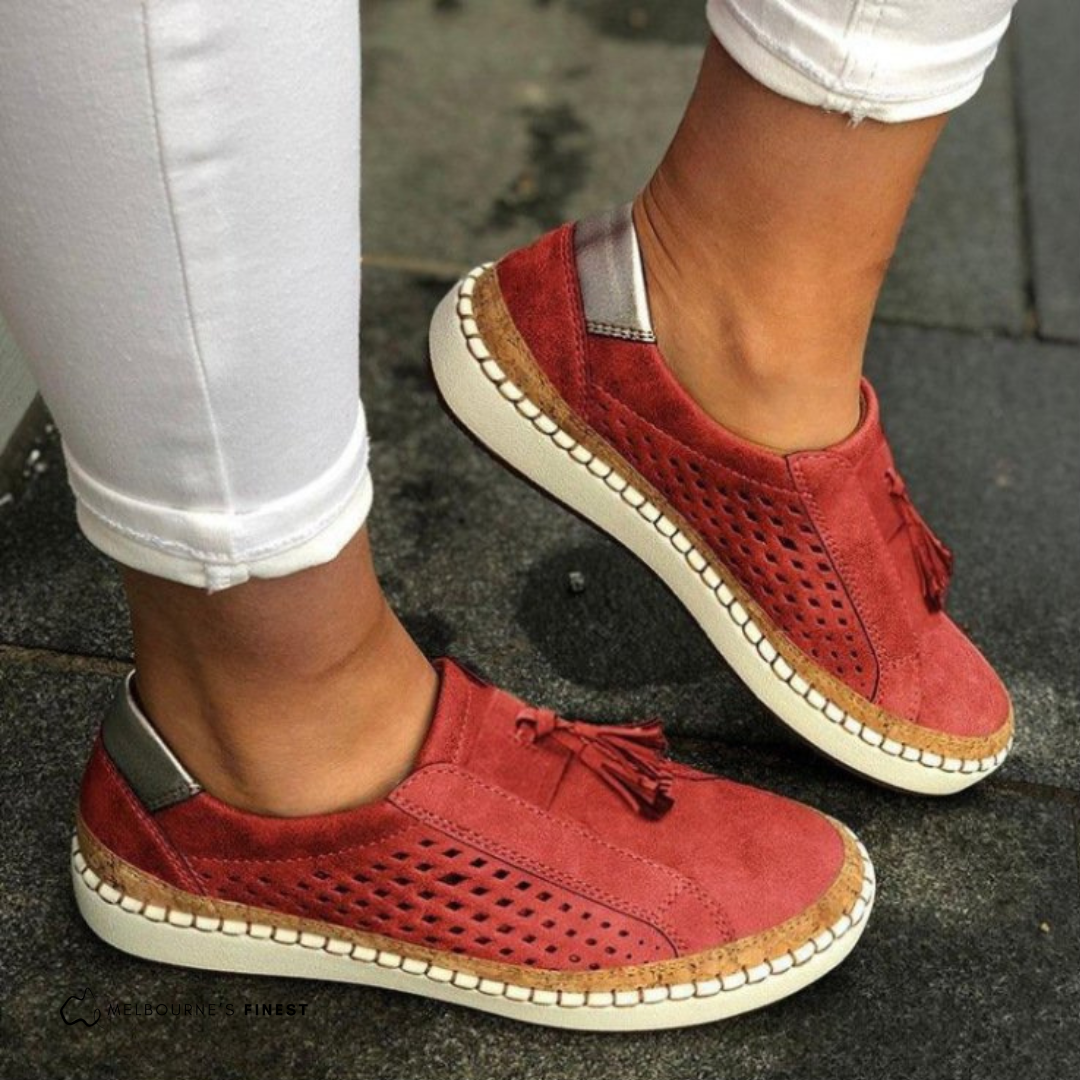 Red perforated slip-on sneakers with front tassel details and contrasting stitched soles, shown from a side angle on a paved surface.