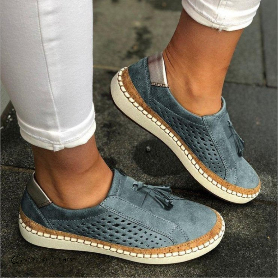 Blue-gray perforated slip-on sneakers with decorative tassels and contrasting stitched soles, shown worn with white cropped jeans.