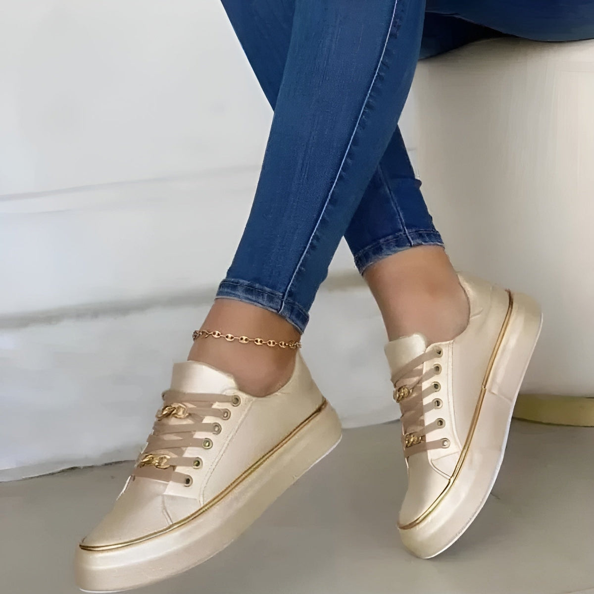 Cream-colored platform sneakers with contrasting gold-tone trim and chain lace details, shown in a crossed-leg side view.