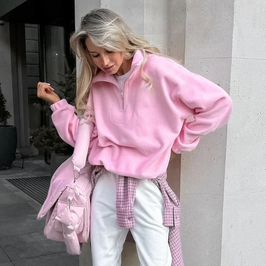 Model styling the pink sweatshirt with the collar partially zipped, gathered sleeves, white trousers, and a pink shoulder bag.