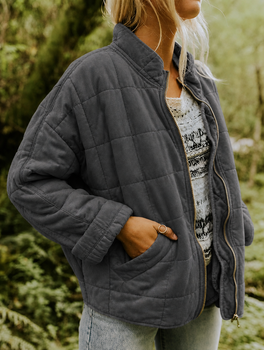 Angled close view of the charcoal jacket showing its raised collar, open front zip, rounded patch pocket, and quilted sleeve.