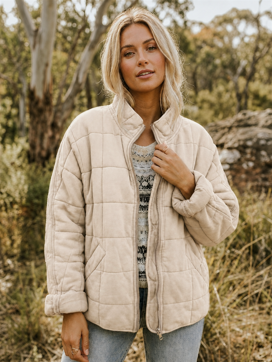 Front view of the jacket in a light neutral shade, featuring a stand collar, geometric quilting, front zip, and patch pockets.