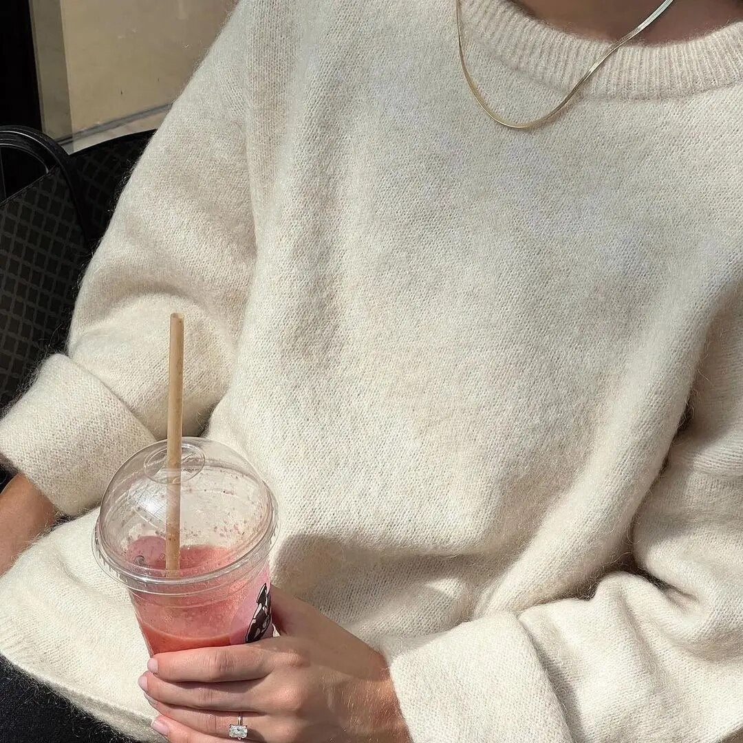 Close view of a cream crew-neck sweater with a necklace and wide folded cuffs while holding a drink.