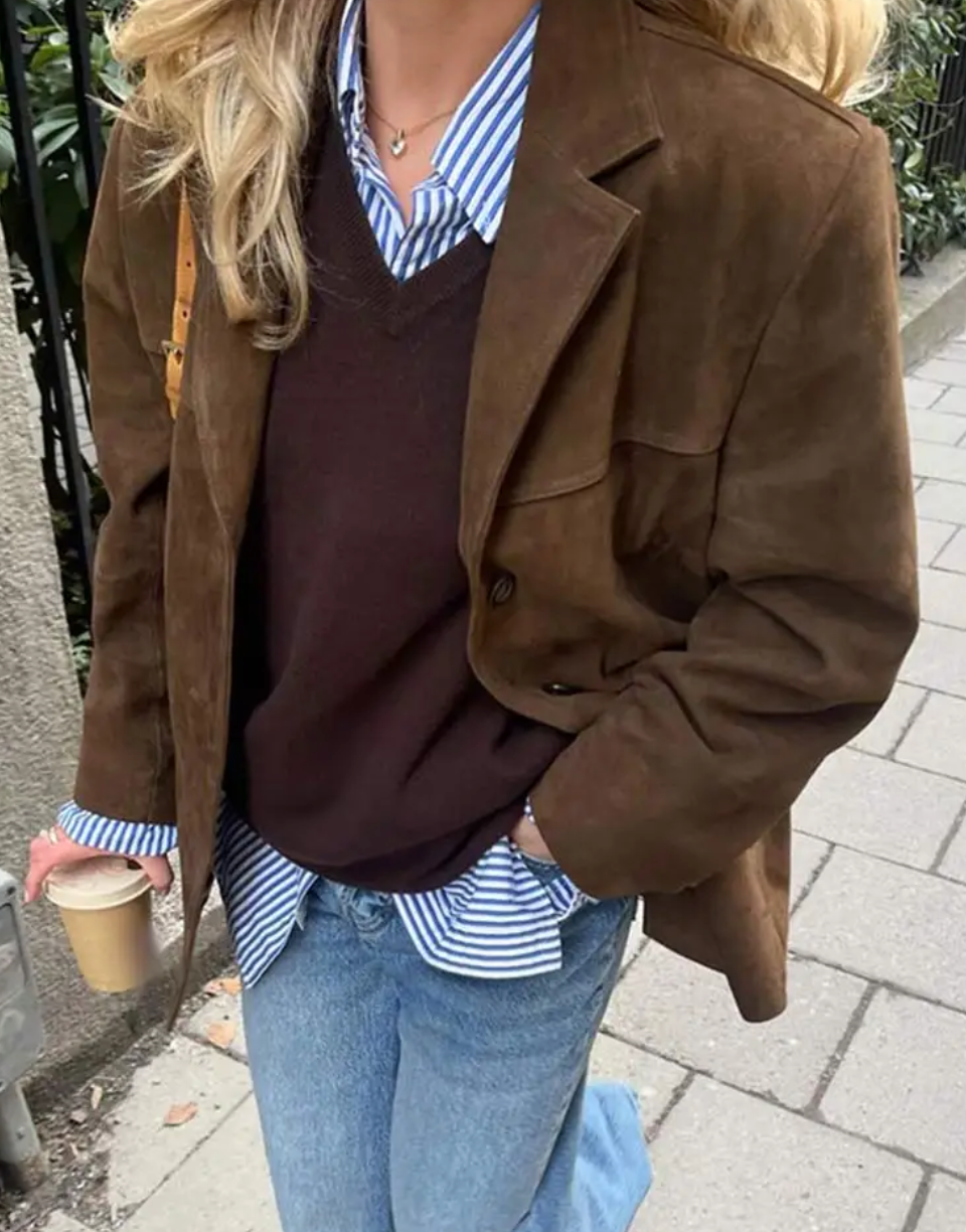Front three-quarter view of the brown blazer worn over layered tops, highlighting the notch lapels, single-button fastening, chest detail, and hand in a front pocket.