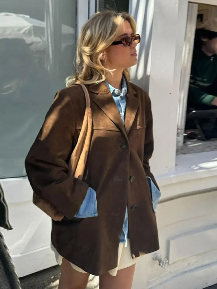 Outdoor side-facing view of the brown relaxed blazer layered over a blue shirt, with its long sleeves, notch lapels, button front, and straight hem visible.