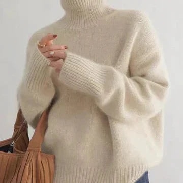 Cream relaxed turtleneck sweater shown from the front with oversized sleeves, ribbed cuffs and hem, styled with a pleated brown handbag.