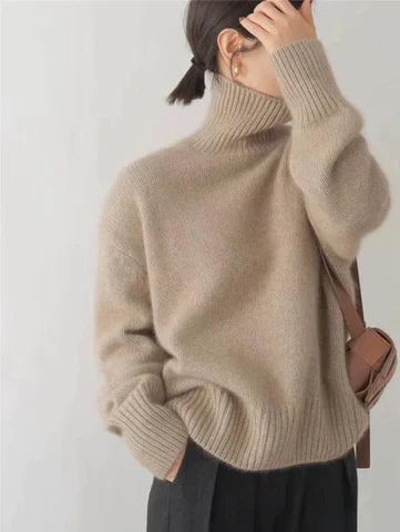 Side view of a beige relaxed turtleneck sweater with a tall ribbed collar, voluminous sleeves and ribbed hem, worn with dark trousers.