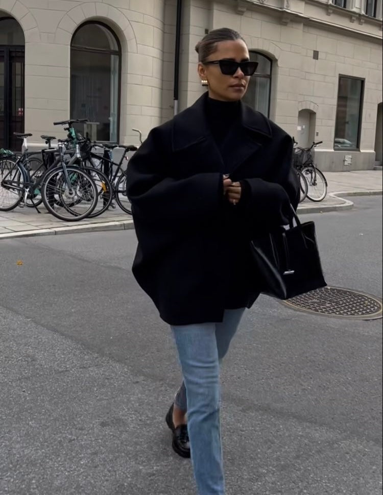 Black short oversized coat worn closed over trousers, showing the broad point collar and relaxed layering fit.