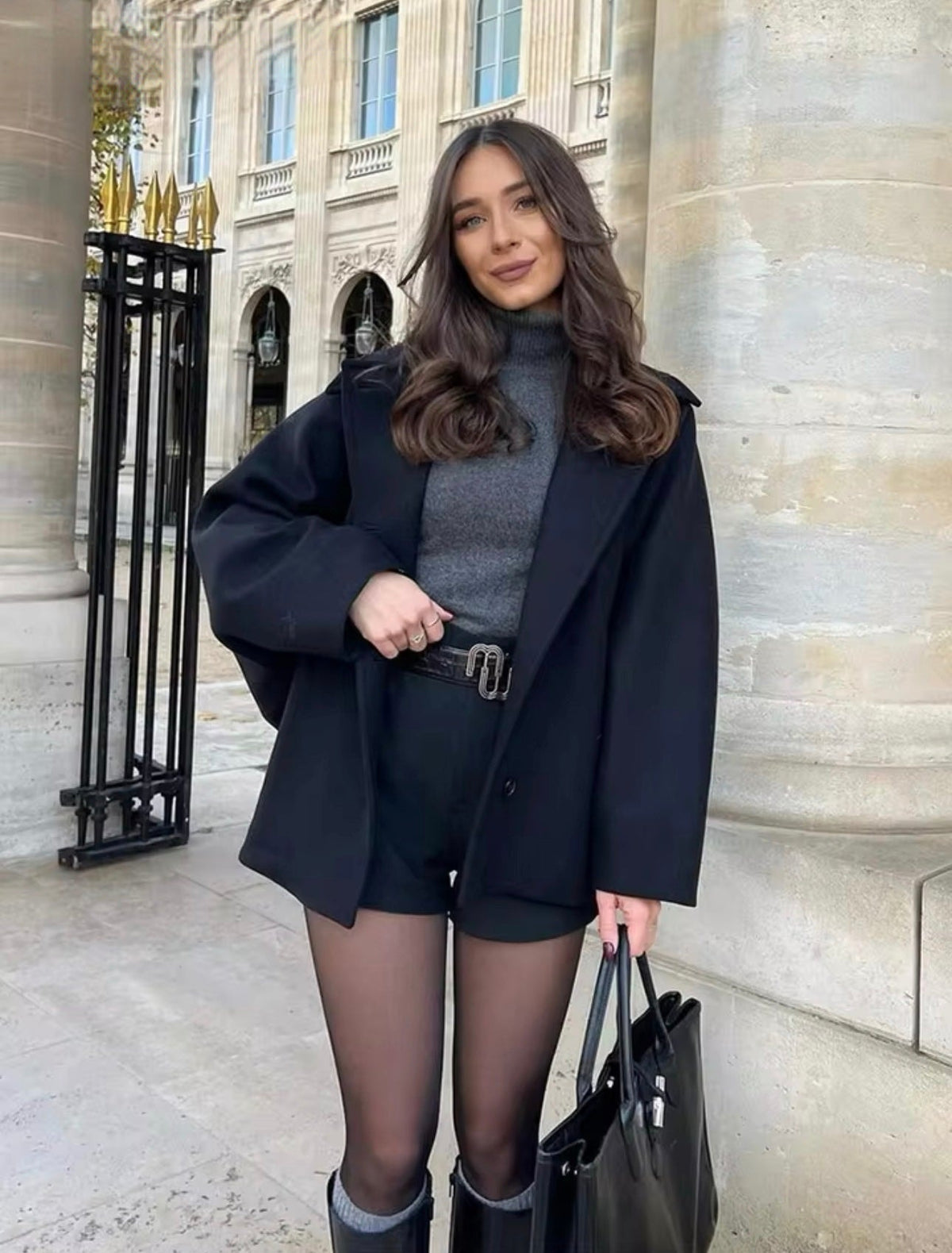Model wearing the black version of the short oversized coat open over a dark outfit, with its point collar visible.