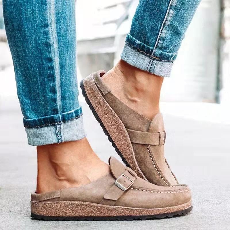 Taupe slip-on flats shown in side profile, highlighting the low textured sole, open heel area, buckle strap, and stitched toe panel.