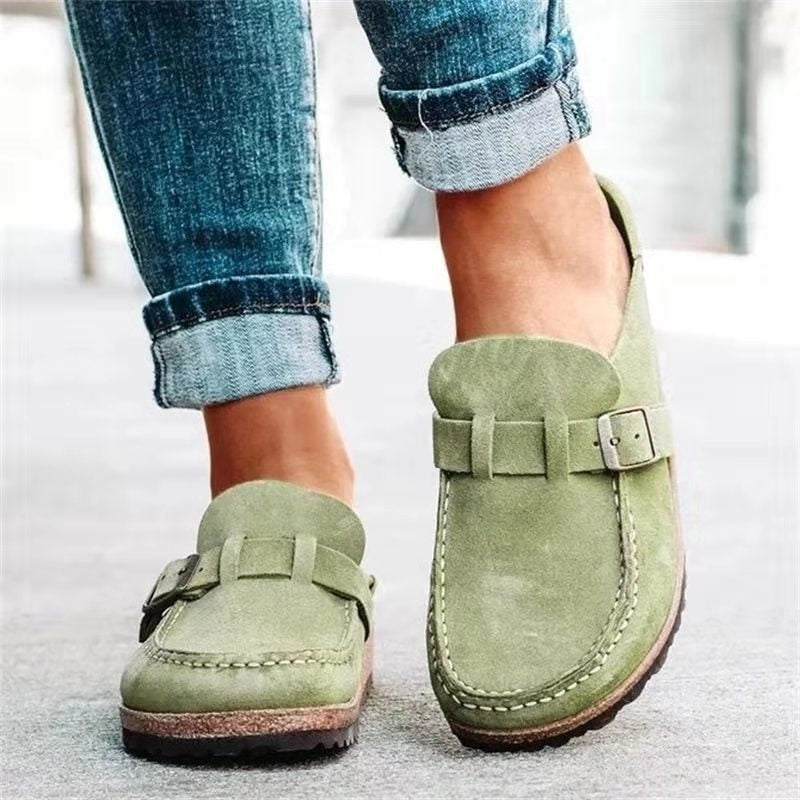 Light sage green slip-on flats with rounded moccasin-style toes, buckle straps, visible stitching, and dark textured soles.
