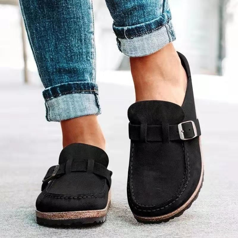 Black slip-on flats with rounded toes and matching buckle straps, shown close up to emphasize the stitched front panels and textured soles.