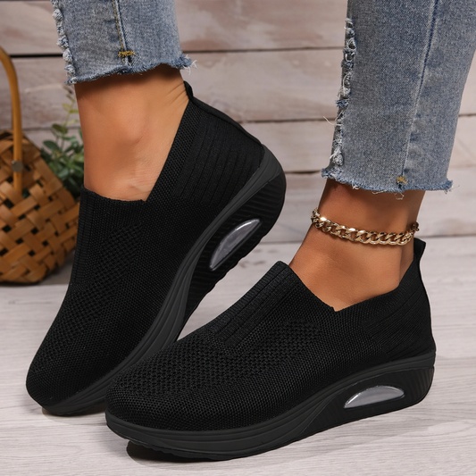 Black slip-on platform walking sneakers shown on feet from a side angle, with textured uppers and oval sole accents.