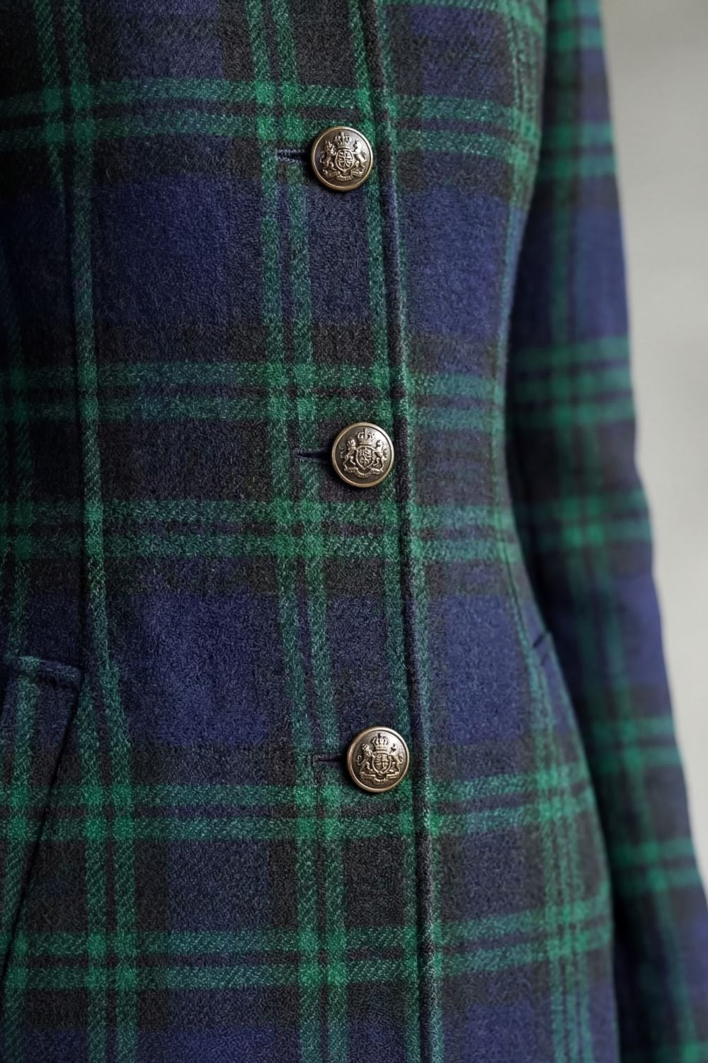 Close-up of the coat’s dark tartan check front with three decorative buttons and a visible pocket opening.