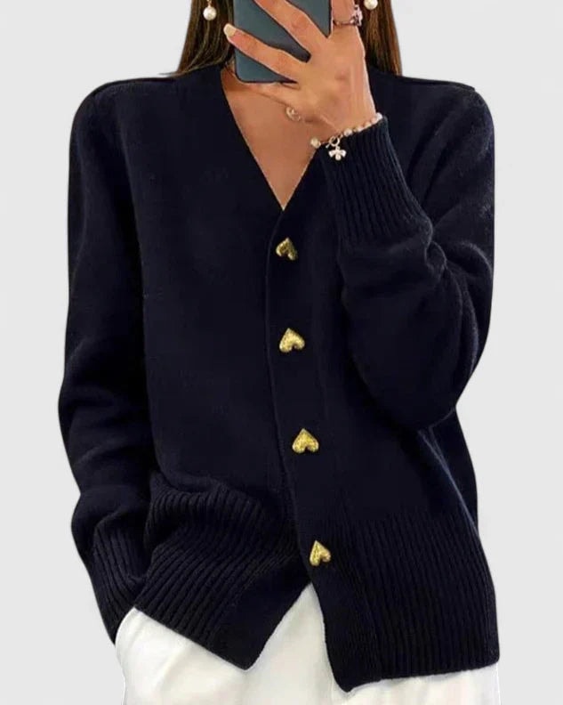 Black V-neck cardigan with gold heart-shaped buttons, long sleeves, and ribbed cuffs and hem, styled over white trousers.