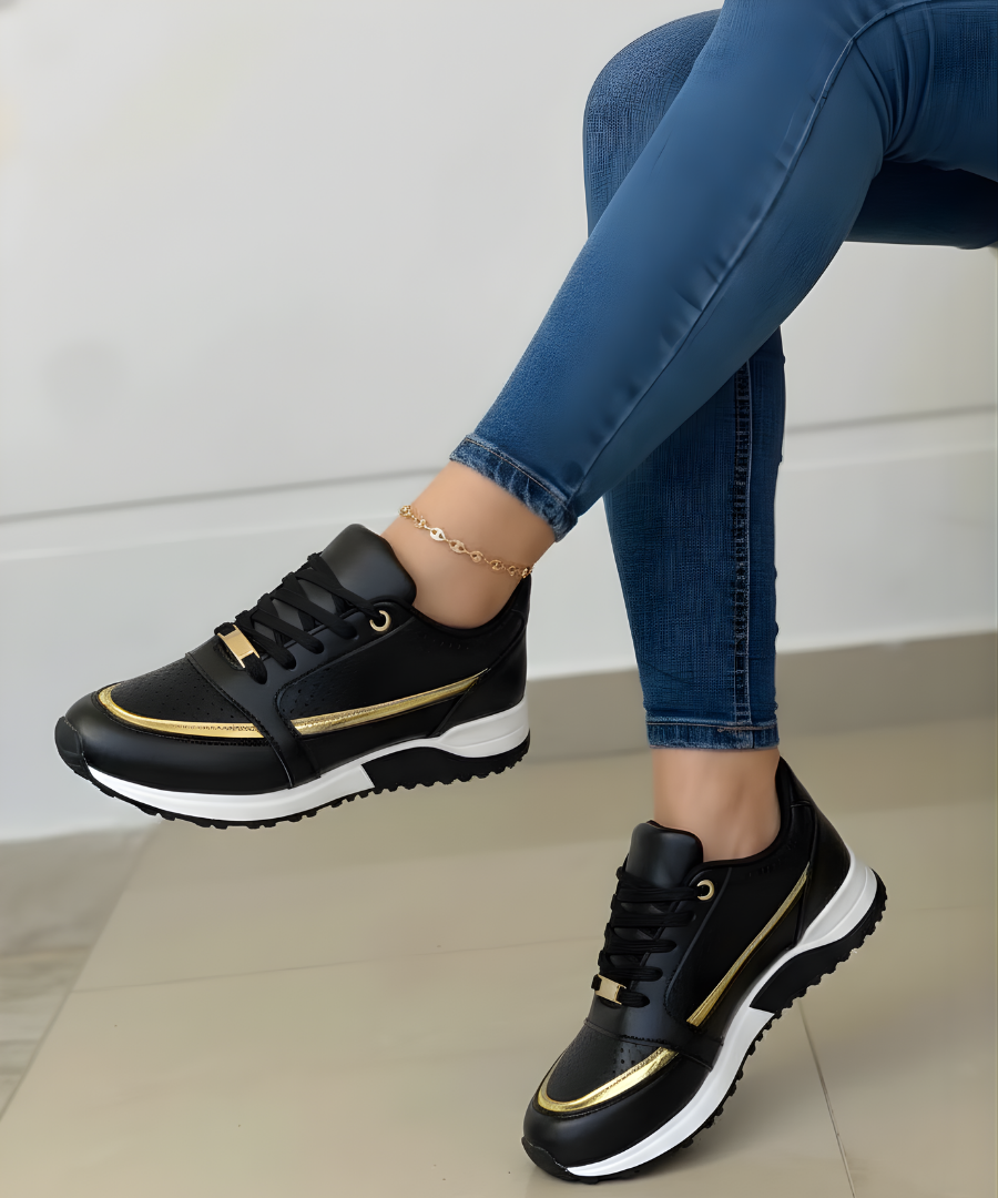 Black low-top women's lace-up sneakers with metallic gold trim and a bold white contrast sole, shown worn with jeans.